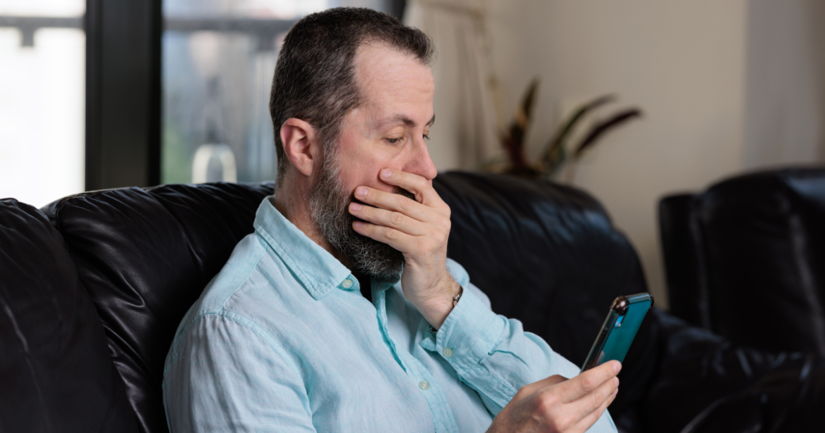 A middle-aged man sitting on a couch, staring at his smartphone in shock and concern. He covers his mouth with one hand, seemingly reacting to something alarming on the phone screen.